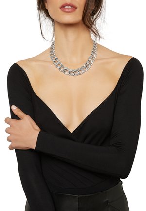 Collana a catena argentata spessa su una modella che indossa un top a maniche lunghe nero con un design a scollatura a V profonda, caratterizzato da una texture liscia e una silhouette aderente.