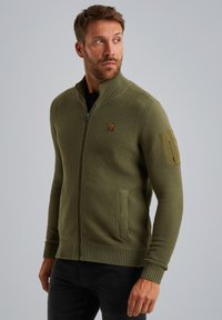 Olivgrüne gestrickte Zip-Jacke mit hohem Kragen, gerippten Bündchen, zwei Seitentaschen und einer aufgesetzten Tasche am linken Ärmel. Auffälliges strukturiertes Muster.