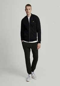 Svart bomberjacka med dragkedja och ribbade ärmslut, kombinerad med mörka byxor och vit t-shirt. Modellen bär grå sneakers. Har en diskret logotyp.