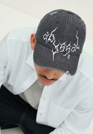 Man wearing a black distressed baseball cap with white lightning-style embroidery, a white button-up shirt, and dark pants.