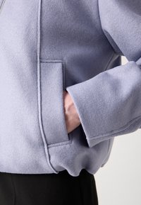La main d'une personne placée dans la poche latérale d'une veste en laine violet clair avec des détails de couture visibles et un large bracelet de manche.