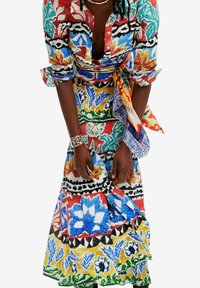 Femme ajustant une longue robe aux motifs floraux colorés et audacieux, avec une écharpe à imprimé mixte vibrant nouée à la taille, portant un bracelet et un collier en argent.