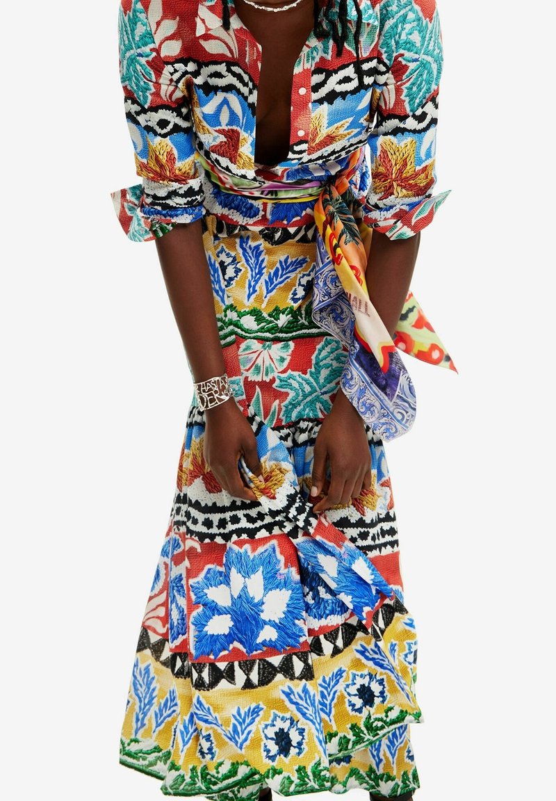 Femme ajustant une longue robe aux motifs floraux colorés et audacieux, avec une écharpe à imprimé mixte vibrant nouée à la taille, portant un bracelet et un collier en argent.