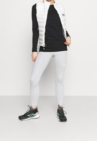 Weiße gepolsterte Weste über einem schwarzen Langarmshirt, kombiniert mit hellgrauen Leggings und schwarzen Sportschuhen mit türkisfarbenen Akzenten.