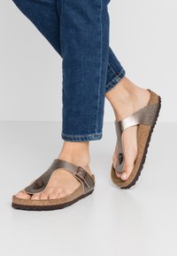 Hnědé kovové flip-flop sandály s texturovanou korkovou stélkou a nastavitelným popruhem, umístěné na pozadí modrých džínů.