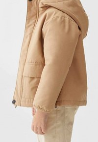 Giacca a vento di color marrone chiaro con cappuccio, polsini elasticizzati e tasca frontale. Tessuto liscio e morbido. Indossata da un bambino.