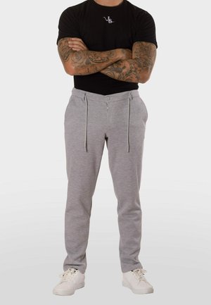 Hombre con los brazos tatuados cruzados, vistiendo una camiseta negra, pantalones gris claro con cordón y zapatillas blancas, de pie frente a un fondo liso.