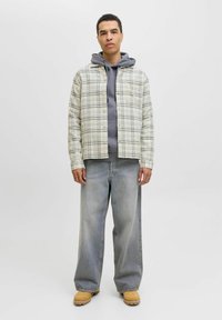 Hemd mit Knopfleiste im hellen Karomuster mit einem grauen Kapuzenpullover darunter. Locker sitzende hellblaue Jeans und braune Stiefel.