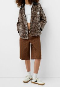 Osoba ubrana w kurtkę w panterkę na białą koszulkę crop, brązowe szorty do kolan, białe skarpetki i białe trampki z gumową podeszwą.