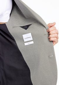 Slim fit-jacka med en texturerad grå exteriör och svart foder. Har en knapp, inre fickor och en tvättrådsetikett synlig inuti.