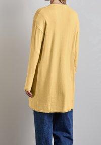 Žlutý pletený top s dlouhými rukávy, volného střihu, kulatým výstřihem a bočními rozparky, kombinovaný s modrými denimovými kalhotami. Měkká textura a minimální švy.