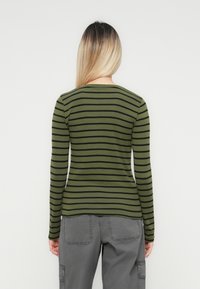 Woman with long blonde ombre hair wearing an olive green and black striped long-sleeve shirt and gray cargo pants, facing away.