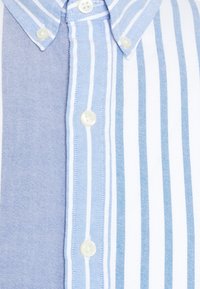 Camicia con bottoni caratterizzata da strisce verticali azzurro chiaro e bianche, realizzata in tessuto morbido con una texture liscia e chiusure a bottoni bianchi.