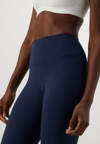 Närbild av en person som bär marinblå högmidjade leggings och en vit crop top, som visar torso, höfter och armar mot en enhetlig bakgrund.
