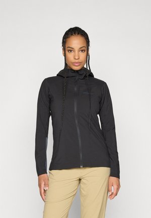 Vrouw met lange vlechten, gekleed in een strakke zwarte hooded jas en beige broek, staande tegen een effen lichte achtergrond.