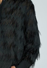 Top negro con un patrón texturizado en zigzag, hecho de un material transparente con largas franjas en los puños y el dobladillo.