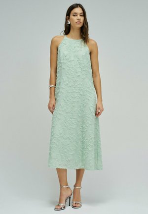 Mujer con vestido midi sin mangas de color verde pálido texturizado y sandalias plateadas de tacón alto, de pie contra un fondo claro y sencillo.