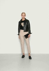 Svart skinnjakke, grafisk svart T-skjorte, beige skreddersydde bukser, svart clutch og loafers, står på et lyst gulv mot en blek vegg.