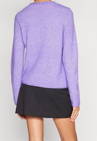 Pull en tricot texturé violet avec un col rond et des manches longues, associé à une jupe noire. L'ourlet du pull tombe à la taille.