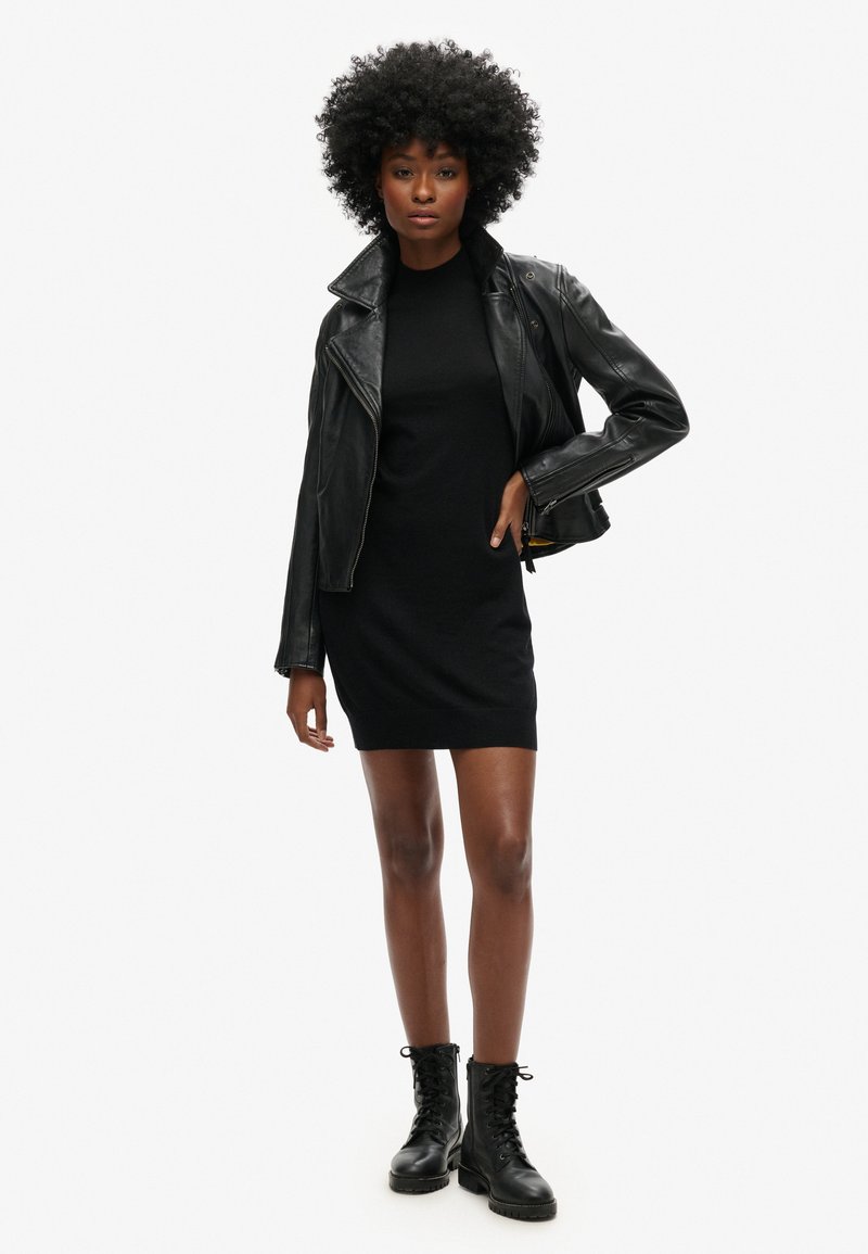 Schwarze Leder-Bikerjacke über einem schwarzen, figurbetonten Kleid, kombiniert mit schwarzen Schnürkampfschuhen; glatte Texturen und schlichte Silhouetten.
