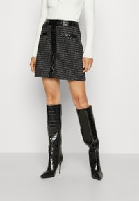 Zwarte tweed mini rok met een gestructureerd patroon, voorzien van een glanzende zwarte leren tailleband, voorknopjes en twee zijzakken.