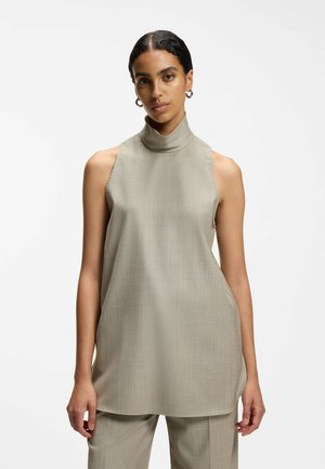 Vrouw met donker haar in een mouwloos beige coltruitje en bijpassende broek staand tegen een witte achtergrond.