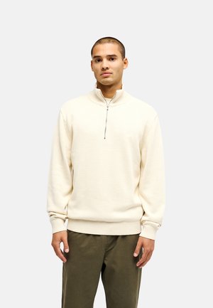 Man standing, wearing a cream half-zip sweater and olive green pants, facing forward against a plain light gray background.