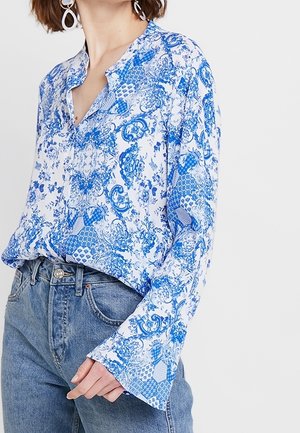 Blouse à motifs bleus et blancs avec un col et des manches évasées, associée à un jean en denim bleu clair. Texture de tissu douce.