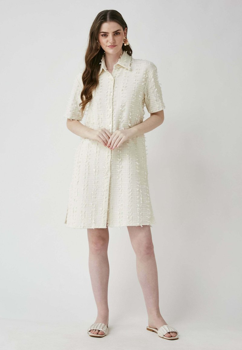 Kurzärmliges beiges Kleid mit strukturiertem Muster, Knopfleiste und Seitenschlitzen. Kombiniert mit flachen, hellen Sandalen.
