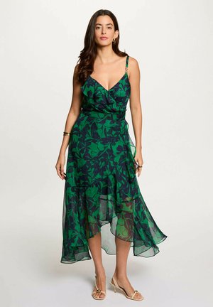Donna che indossa un vestito midi floreale verde e blu navy senza maniche con orlo arricciato e tacchi aperti beige, in piedi davanti a uno sfondo semplice.