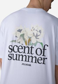 Hvid bomulds T-shirt med et stort blomsterdesign med hvide blomster og grønne accenter samt teksten "duft af sommer" i fede sorte bogstaver.