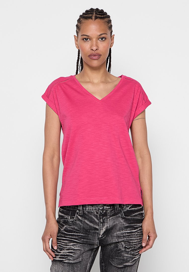 Rosa, kurzärmliges V-Ausschnitt-T-Shirt mit weichem Textil und kleinen seitlichen Schlitzen. Kombiniert mit grauen, verwaschenen Jeans.