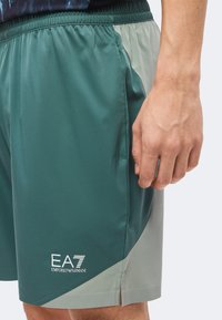 Άνδρας που φοράει πράσινα και γκρι σορτς EA7 Emporio Armani, κοντινή λήψη στην πλαϊνή τσέπη και το χέρι που ακουμπά δίπλα της.