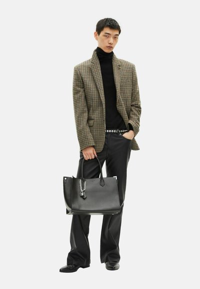 Bruine geruite blazer, zwarte coltrui, zwarte broek en riem. Hij houdt een grote zwarte leren tote met kettingdetail en platte handvatten vast.