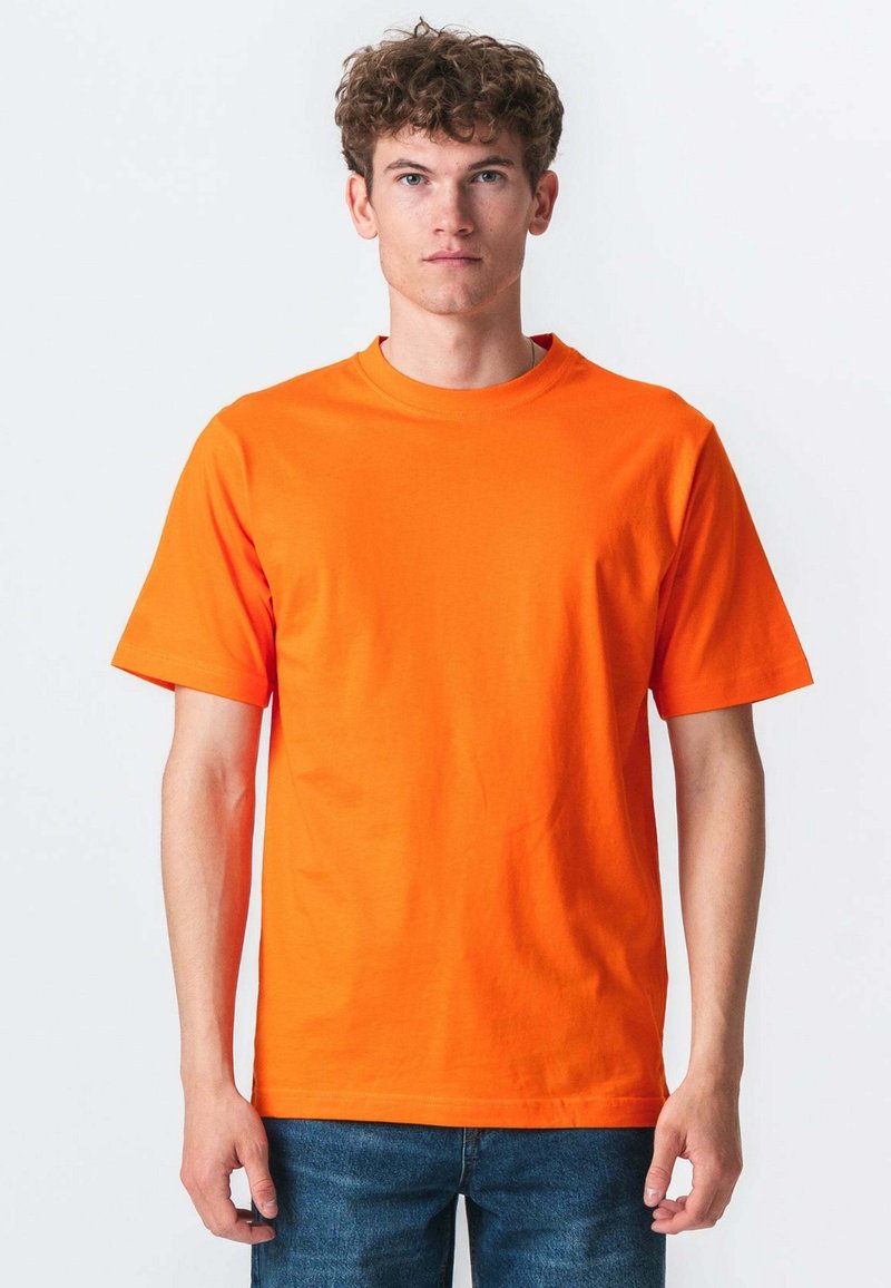 Ung mand med krøllet hår iført en ensfarvet lys orange kortærmet t-shirt og blå jeans mod en hvid baggrund.