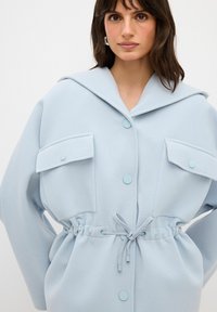 Camisa overshirt de color azul claro con una gran capucha, cierre de botones, cintura elástica y dos bolsillos frontales. Hecha de un tejido suave y texturizado.