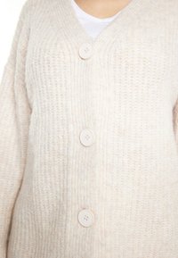 Cardigan beige in maglia con scollo a V, tre grandi bottoni e texture a costine. Materiale morbido con un aspetto accogliente.