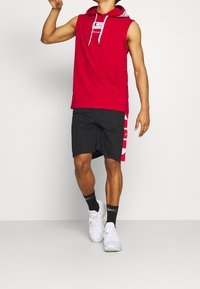 Punainen hihaton huppari, jossa on hupulliset narut, mustat shortsit punaisella sivupaneelilla ja valkoiset urheilukengät. Yksinkertainen malli ja rento istuvuus.