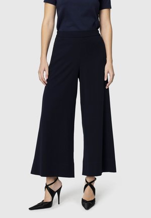 Frau trägt schwarze weit geschnittene, gekürzte Hosen und schwarze High-Heels mit gekreuzten Riemen, steht vor einem einfarbigen Hintergrund.