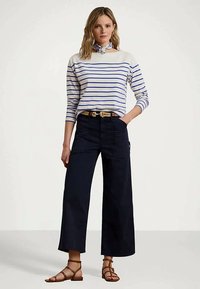 Mulher em pé a usar uma camisa de manga longa branca e azul às riscas, calças largas azul-marinho, sandálias castanhas e um lenço azul atado ao pescoço.