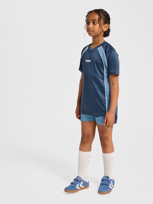 Młoda dziewczyna stojąca w niebieskim stroju sportowym, białych podkolanówkach i niebieskich trampkach na jednolitym jasnym tle.