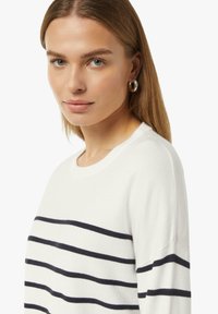 Witte trui met marineblauwe horizontale strepen, ronde halslijn en lange mouwen. Glad textuur en een ontspannen pasvorm. Zilveren ringoorbellen accentueren de look.