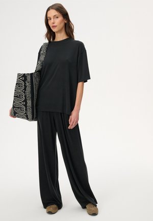 Kobieta w luźnej czarnej bluzce i spodniach, w beżowych tenisówkach, trzyma dużą czarno-białą torbę na prostym tle.