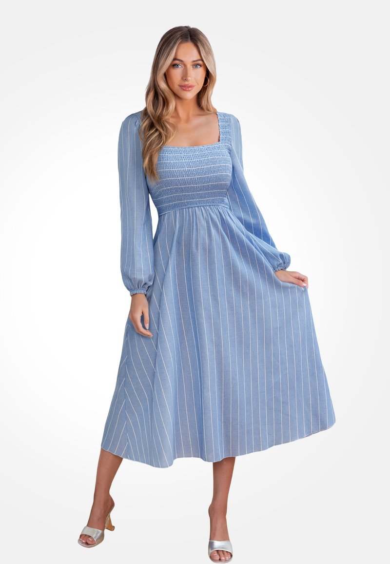 Vestido azul claro con rayas blancas verticales, escote cuadrado, mangas largas y corsé fruncido. Llega hasta la mitad de la pantorrilla. Sandalias de tacón plateadas.