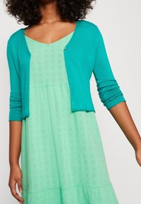 Tyrkysový cardigan s dlouhými rukávy, kombinovaný s texturovanými, světle zelenými šaty se výstřihem do V a rozšířeným lemem.