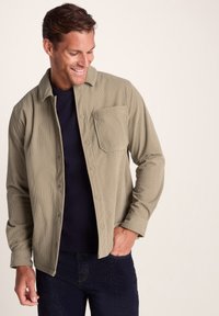 Smiling man in beige corduroy button-up jacket over a black shirt, with one hand in dark blue jeans pocket, standing against a plain background.