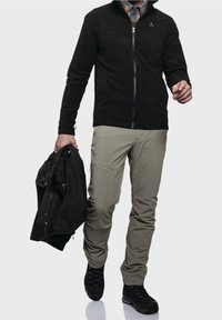 Muž v černé bundě na zip, béžových cargo kalhotách a černých turistických botách, drží v levé ruce černou bundu a kráčí vpřed.