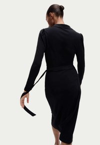 Vestido negro envolvente con mangas largas y cuello, que presenta hombros fruncidos y un cinturón atado en la cintura. Tejido suave con una silueta ajustada.