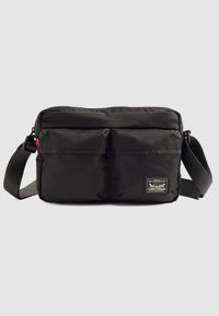 Borsa a spalla nera Levi's con due tasche frontali, tracolla regolabile e un'etichetta con il logo su uno sfondo grigio chiaro uniforme.