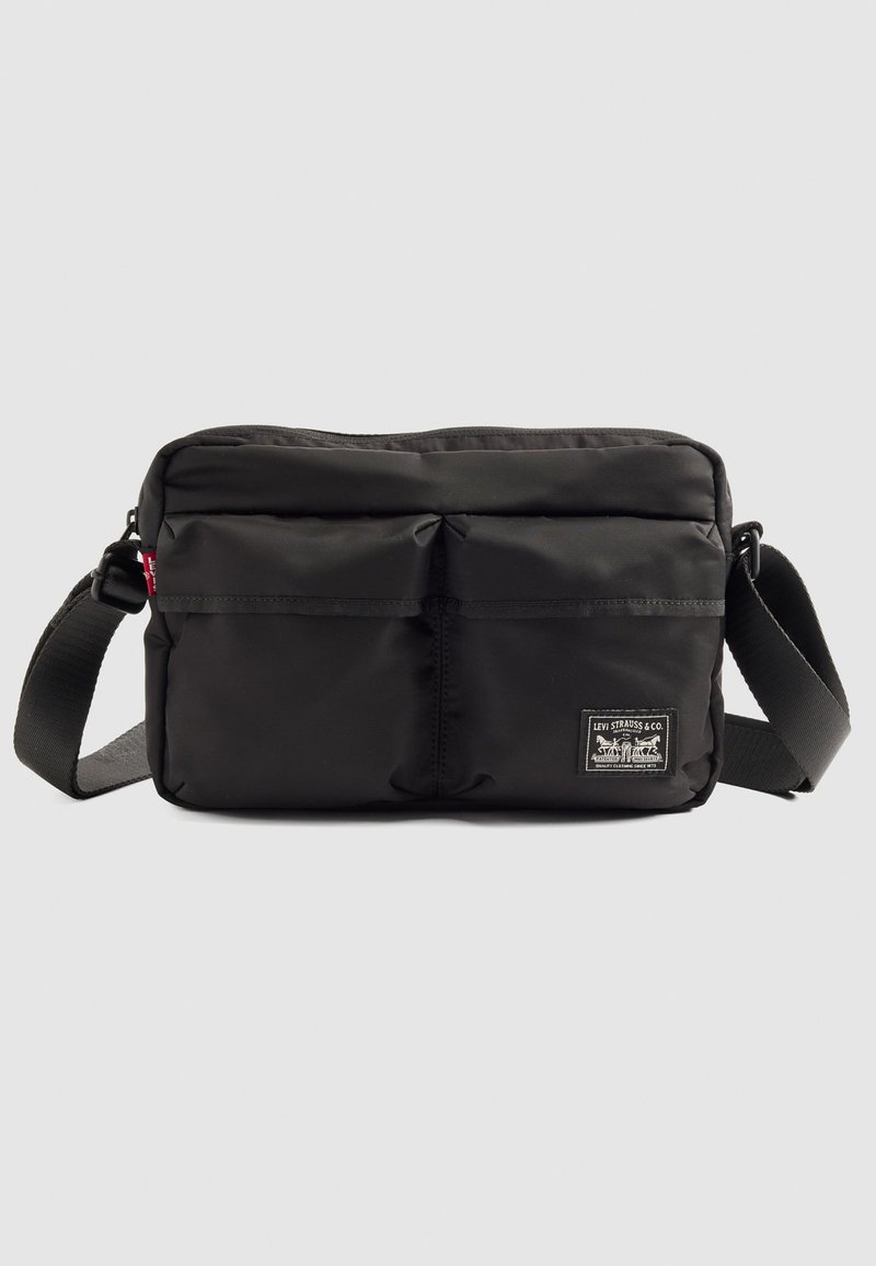 Borsa a spalla nera Levi's con due tasche frontali, tracolla regolabile e un'etichetta con il logo su uno sfondo grigio chiaro uniforme.